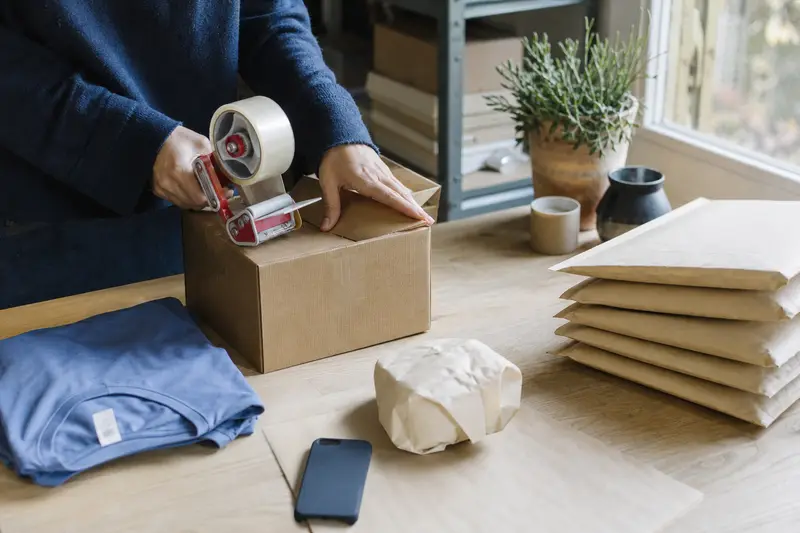 Hands taping up a parcel on a packing bench beside a folded T-shirt, a wrapped mug, a phone case and padded mailers