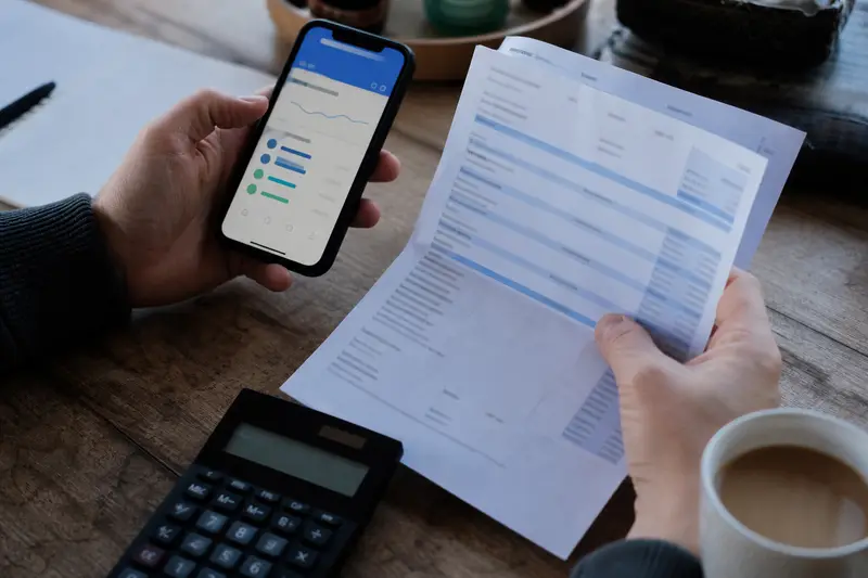 Hands holding a bank statement and a phone banking app over a desk with a calculator and a mug of tea