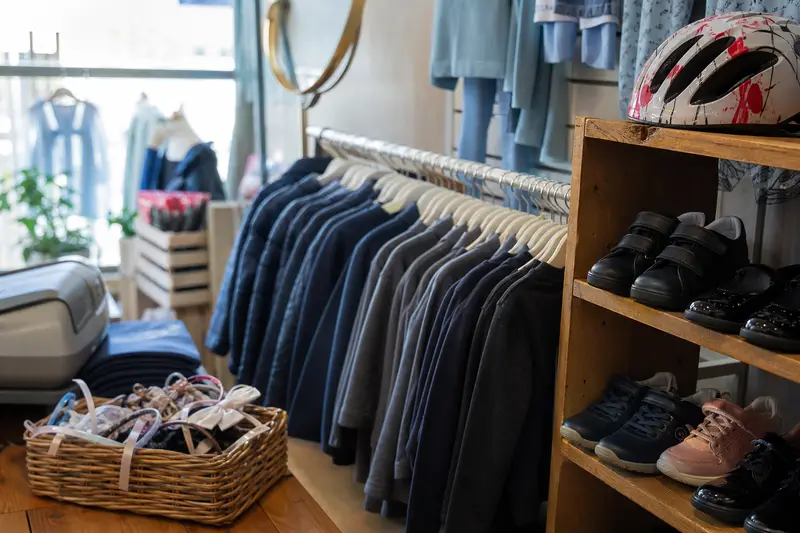 A children's clothes shop with a rail of navy and grey school jumpers, a shelf of children's school shoes and trainers, a cycle helmet and a basket of hair accessories