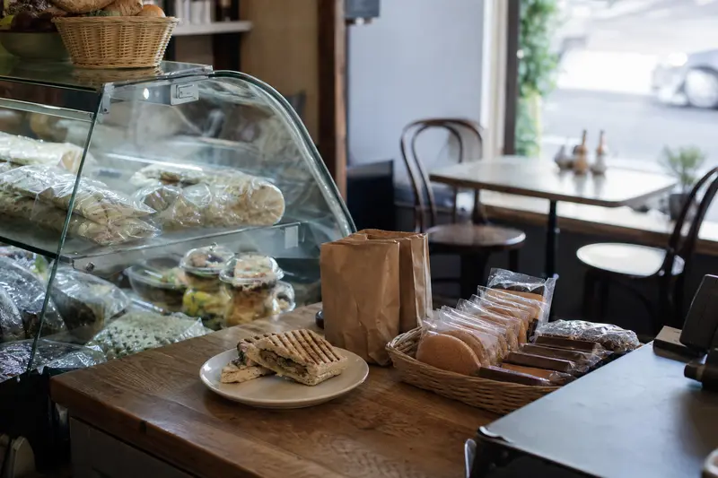 A café counter with wrapped sandwiches in a chiller, a toasted panini on a plate, paper takeaway bags and a basket of biscuits by the till